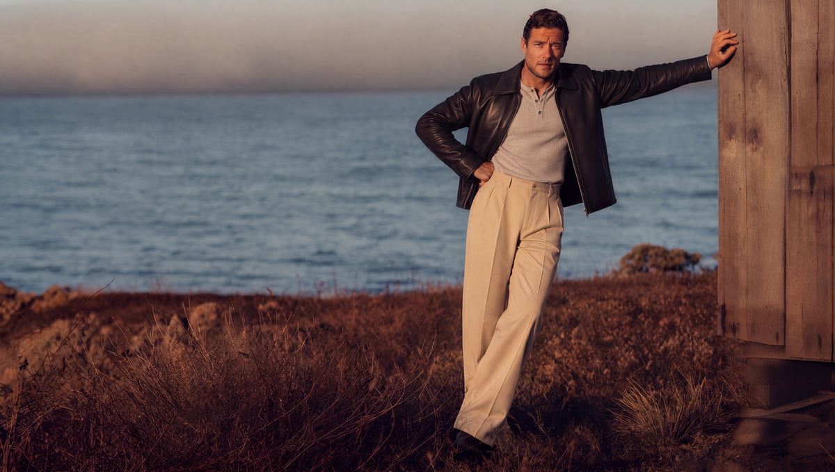 bydefault. black leather jacket and beige tailored trousers, coastal cliff at sunset