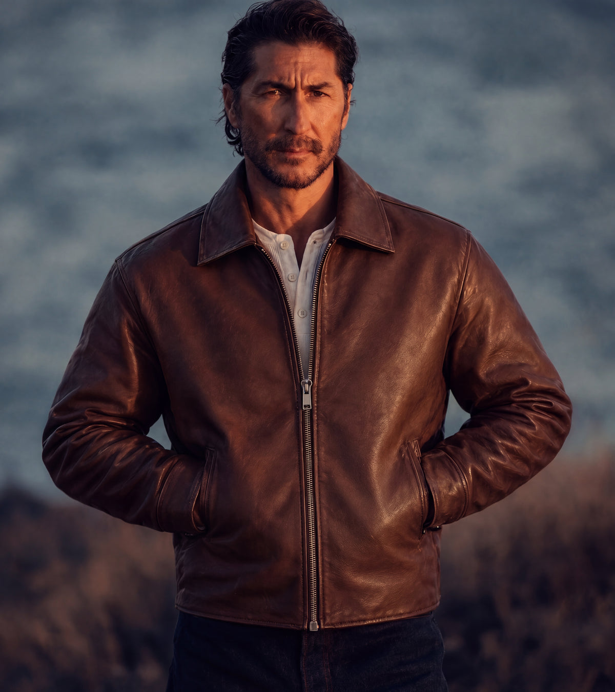 bydefault. brown leather jacket and white henley, coast at dusk